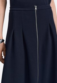 Meriinisinine A-lõikelise seelik tekstuurse kangaga, mille keskel on hõbedane lukk ja voldid, mis lisavad liikumist ja vormi.