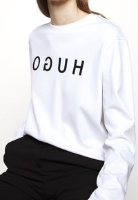 Camisa blanca de manga larga con el texto negro "HUGO" impreso al revés en la parte frontal. Hecha de una tela de algodón suave. Combinada con pantalones negros.