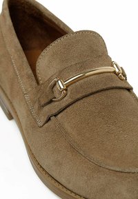 Pruunist nahast loafers, millel on kuldtooni metallist aksent, katkematu õmblus ja madal profil. Kujundus on klassikaline ja funktsionaalne.