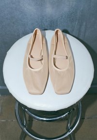 Paire de ballerines en mesh beige à bouts carrés et brides simples, posées sur un tabouret rond blanc rembourré avec des pieds en métal.