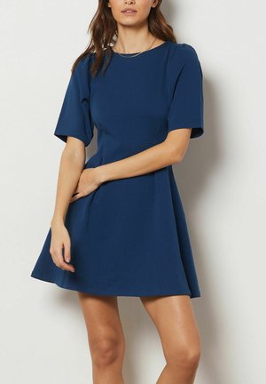 Femme portant une robe bleu marine à manches courtes, longueur genoux, debout contre un mur blanc avec une main reposant sur l'autre bras.