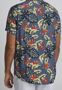 Korte mouwen shirt met een blauwe achtergrond en grote tropische bladmotieven in groen, geel en rood, gemaakt van lichte stof.