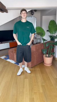Green short-sleeve t-shirt, black shorts, and white sneakers worn by a person standing indoors. Furniture and large potted plant in background.
