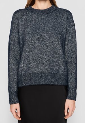 Personne portant un pull en maille texturée bleu marine à manches longues avec des poignets côtelés, associé à une jupe noire.