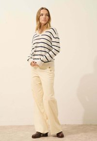 Sweter w paski w kolorze kremowym i granatowym, w luźnym kroju, zestawiony z jasnymi żółtymi spodniami z sztruksu w fasonie dzwonów z guzikami, noszonymi z brązowymi, grubymi obcasami.