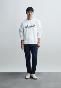 Sweatshirt blanc avec un texte "Dsquared²" en cursive noir, associé à un jean bleu foncé et des baskets crème. Le modèle pose devant un fond neutre.