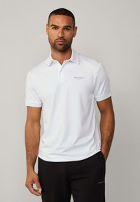 Weißes Poloshirt aus glattem Stoff mit spitzem Kragen. Mit einem Logo auf der linken Brust. Kombination mit schwarzen Hosen für einen Kontrast.