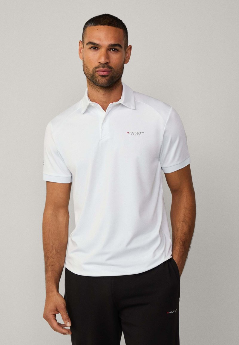 Weißes Poloshirt aus glattem Stoff mit spitzem Kragen. Mit einem Logo auf der linken Brust. Kombination mit schwarzen Hosen für einen Kontrast.