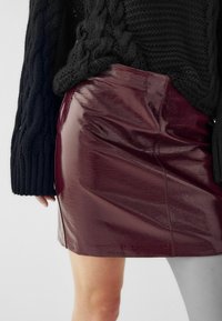 Bordo umetni usnjeni mini krilo z lesketajočo površino, ki ima gladko teksturo in podrobnosti na stranskih šivih, v kombinaciji s črnim puloverjem z pleteno teksturo.