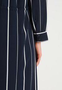 Mörkblå klänning med vertikala vita ränder, med en knäppning vid ärmslutet och en bälte i midjan. Materialet ser slätt och något strukturerat ut.