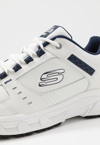 Sneaker bianco con accenti blu navy, caratterizzata da un pannello testurizzato e un design cucito. I lacci sono bianchi con una linguetta in rete e dettagli con logo.