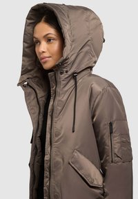 Ruskea topattu takki, jossa on suuri huppu, etuvetoketju ja nepparikiinnitys. Takissa on sivutaskut ja lisävetoketjutasku hihassa.