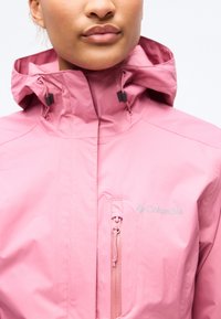 Gros plan sur une personne portant une veste Columbia rose avec une capuche, un col haut et une poche poitrine à fermeture éclair verticale.