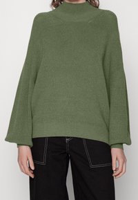 Zielony, oversize'owy sweter z wysokim dekoltem, prążkowaną teksturą i szerokimi rękawami. Do tego czarne spodnie z białym przeszyciem.