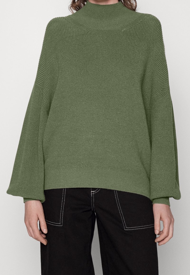 Zielony, oversize'owy sweter z wysokim dekoltem, prążkowaną teksturą i szerokimi rękawami. Do tego czarne spodnie z białym przeszyciem.