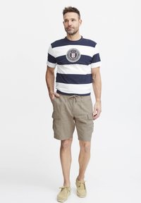 Menns stripete t-skjorte i marineblått og hvitt med en rund logo, kombinert med khaki cargo shorts med lommer. Skoene er beige loafers.