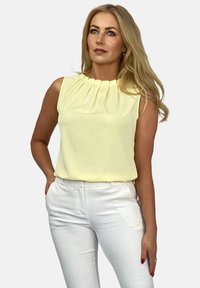 Bez rękawów żółta bluzka z plisowanym dekoltem i gładką teksturą, w zestawieniu z dopasowanymi białymi spodniami z bocznymi kieszeniami.