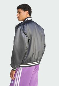 Black satin bomber jacket with white striped collar and cuffs, featuring a relaxed fit and elasticized hem. Paired with purple track pants.