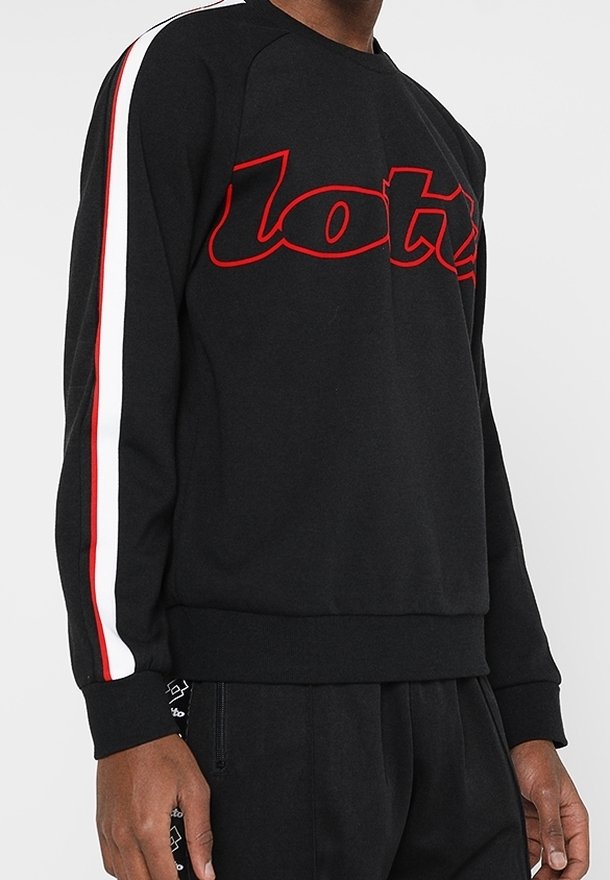 Personne portant un sweat-shirt noir avec un texte contour rouge et des bandes blanches et rouges le long des manches, associé à un pantalon noir.