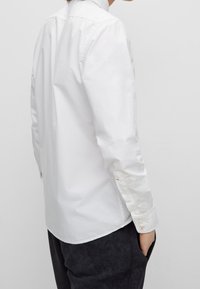 Vit bomullsskjorta med klassisk krage, långa ärmar med knäppta manschetter och rundad nederkant, bärs över mörka byxor.