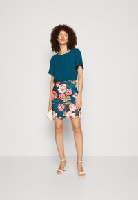Türkise Bluse mit kurzen Ärmeln, kombiniert mit einem floralen Minirock, der pinke Rosen auf blauem Hintergrund zeigt. Das Model hält eine kleine Clutch.