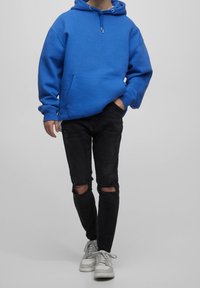 Osoba v modré mikině s kapucí, černých roztrhaných skinny džínách a bílých teniskách stojící na šedém pozadí.