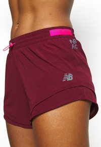 Pantalones cortos atléticos burdeos con un tejido texturizado, cintura elástica, cordón rosa y logo reflectante de New Balance en el lateral.