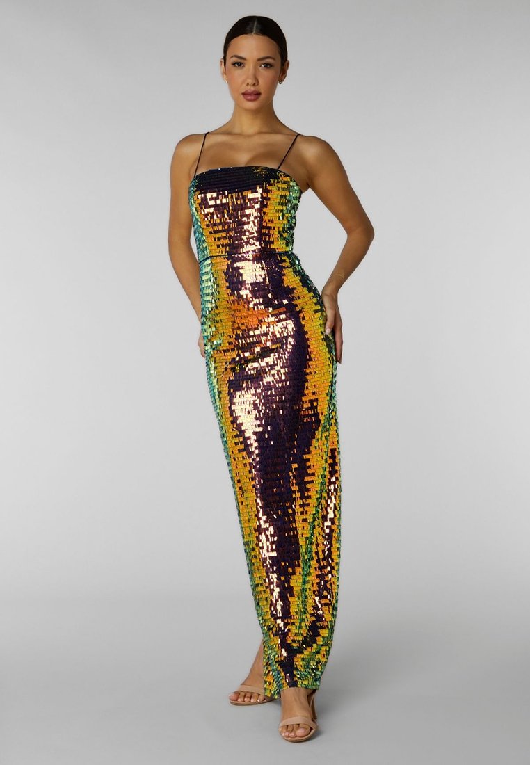 Frau in einem bodenlangen, mehrfarbigen Paillettenkleid mit dünnen Trägern, die mit einer Hand auf der Hüfte vor einem schlichten Hintergrund posiert.