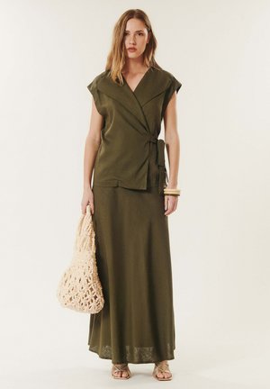 Femme portant un top cache-cœur sans manches vert olive et une longue jupe, tenant un sac cabas tricoté crème, et portant des sandales beiges et des bracelets.