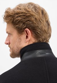 Schwarzer Mantel aus strukturiertem Wollstoff, mit hohem Kragen und einem Lederakzent am Nacken, auf dem der Markenname "DIGEL" geprägt ist.