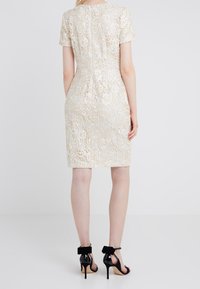 Žena v krémových krajkových šatech po kolena s krátkým rukávem a květinovými vzory, doplněná černými lodičkami na vysokém podpatku s mašličkami.