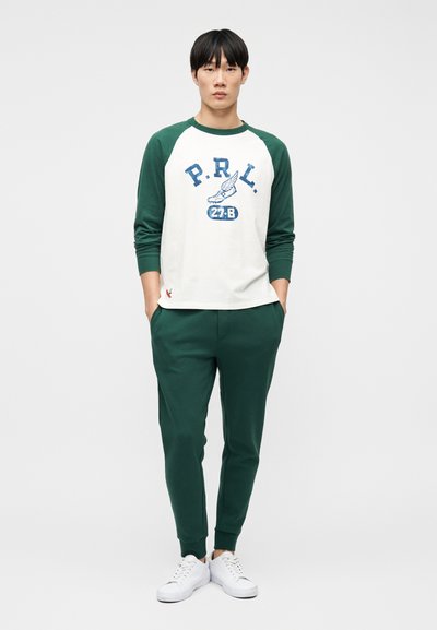 Νεαρός άνδρας που στέκεται φορώντας λευκό και πράσινο μακρυμάνικο πουκάμισο με κείμενο "P.R.L.", πράσινες φόρμες και λευκά αθλητικά παπούτσια, σε λευκό φόντο.