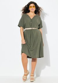 Oliwkowa sukienka z krótkim rękawem w kształcie litery V, z detalami guzików i beżowym paskiem, zestawiona z brązowymi platformami na jasnym tle.