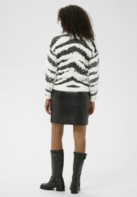 Göndör hajú személy fekete-fehér csíkozott bolyhos pulóverben, fekete bőr miniszoknyában és térdig érő fekete csizmában áll, háttal.