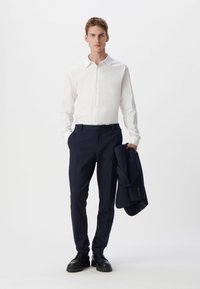 Biała koszula zapinana na guziki, ciemnoniebieskie spodnie, czarne buty, trzymający granatową marynarkę. Minimalistyczny design, dopasowany krój, gładka tekstura.