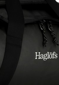 Borsa a tracolla nera realizzata in materiale resistente, con chiusura a cordino e spallacci intrecciati. Il logo "Haglöfs" in bianco è visibile.