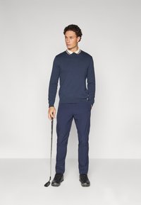 Pull marin avec un col blanc et bordeaux, associé à un pantalon marine et des chaussures de golf noires, tenant un club de golf. Texture de tissu lisse.
