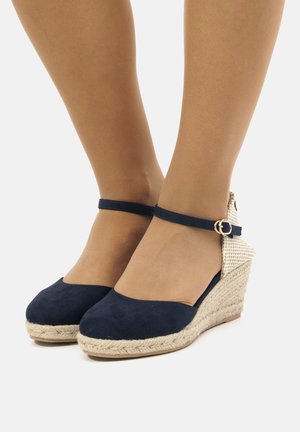 Zapatos de cuña de ante azul marino con un tacón trenzado, correa al tobillo y hebilla dorada. Textura suave con un diseño de punta redondeada.