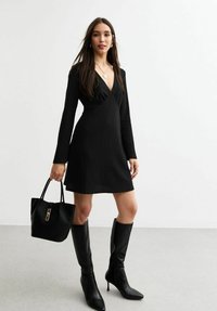 Robe noire à côtes avec des manches longues et un col en V. Portée avec des bottes noires jusqu'aux genoux et un sac à main noir avec quincaillerie dorée.