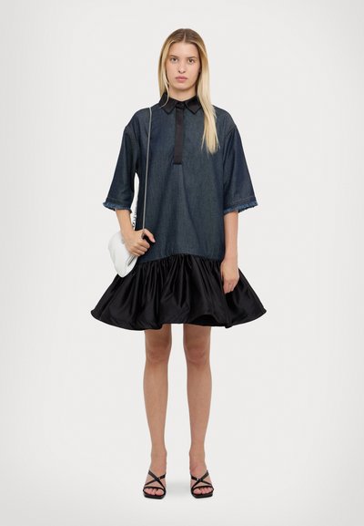 Denim-paita mekko mustalla satiinirikotetulla hameella. Ominaisuuksia ovat napitettu kaulus, lyhyet hihat ja kuluneet hihansuut. Asusteena valkoinen laukku.