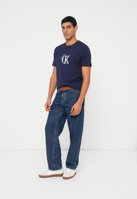 Tumši zils kokvilnas T-krekls ar baltu logotipu, kopā ar vaļīgām zilām džinsa biksēm un baltām sporta kurpēm ar brūnām zolēm. Redzama vienkārša jostas detaļa.