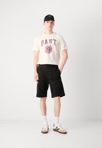 Cream t-shirt with "GANT" graphic, black shorts, white socks, and brown-accented sneakers. Casual outfit on a neutral background.
