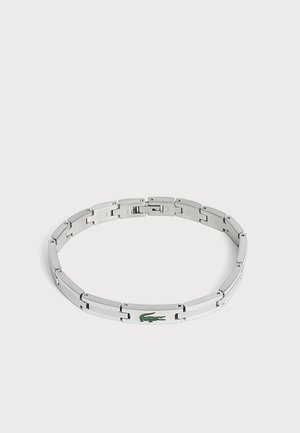 Bracelet en métal argenté avec un logo crocodile vert sur un maillon, présenté sur un fond clair uni.