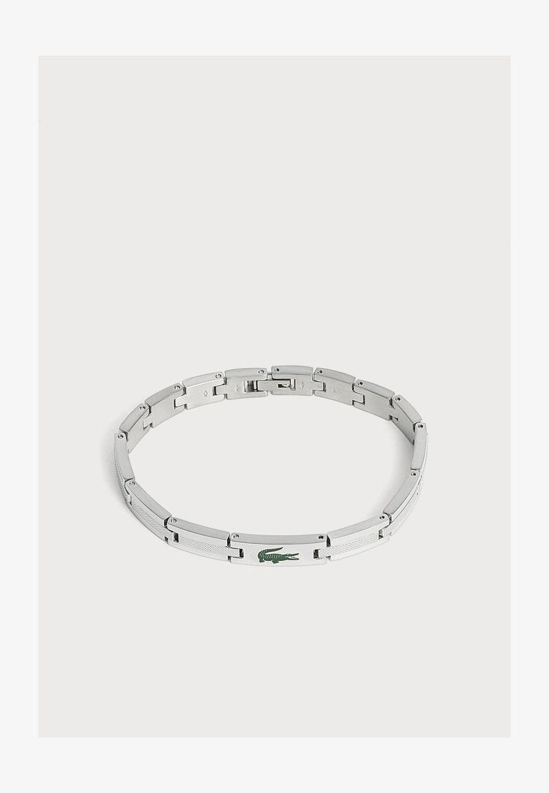 Bracelet en métal argenté avec un logo crocodile vert sur un maillon, présenté sur un fond clair uni.