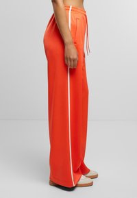 Weit geschnittene Jogginghosen aus leuchtend orangefarbenem Stoff mit einem weißen Seitenstreifen, elastischem Bund mit Kordelzug und seitlichen Taschen.