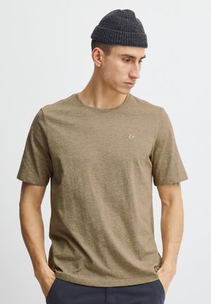 Mann trägt ein braunes kurzärmeliges Hemd und eine dunkelgraue gestrickte Mütze, schaut nach rechts unten mit den Händen in den Taschen, vor einem weißen Hintergrund.