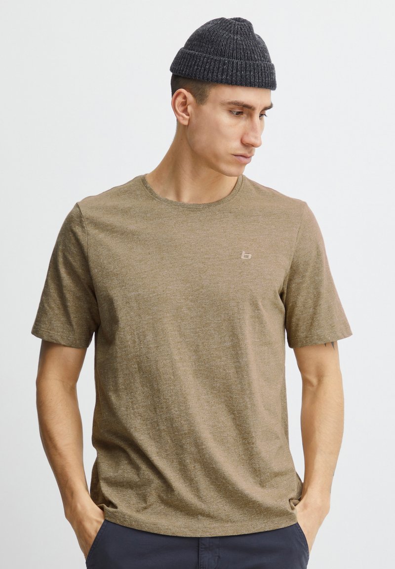 Άνδρας που φορά καφέ κοντομάνικο πουκάμισο και σκούφο πλεκτό σκούρο γκρι, κοιτάζοντας προς τα κάτω στα δεξιά του με τα χέρια στις τσέπες, μπροστά σε λευκό φόντο.