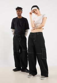Black cargo pants with white stitching, featuring multiple pockets. Complemented by a fitted light pink top and black graphic t-shirt.