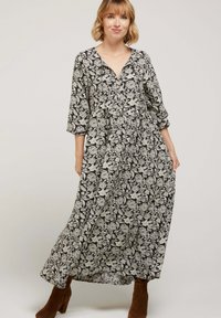 Robe maxi noire avec motif floral blanc, décolleté en V, manches trois-quarts et coupe loose, fabriquée en tissu léger. Bottes à talons brunes.