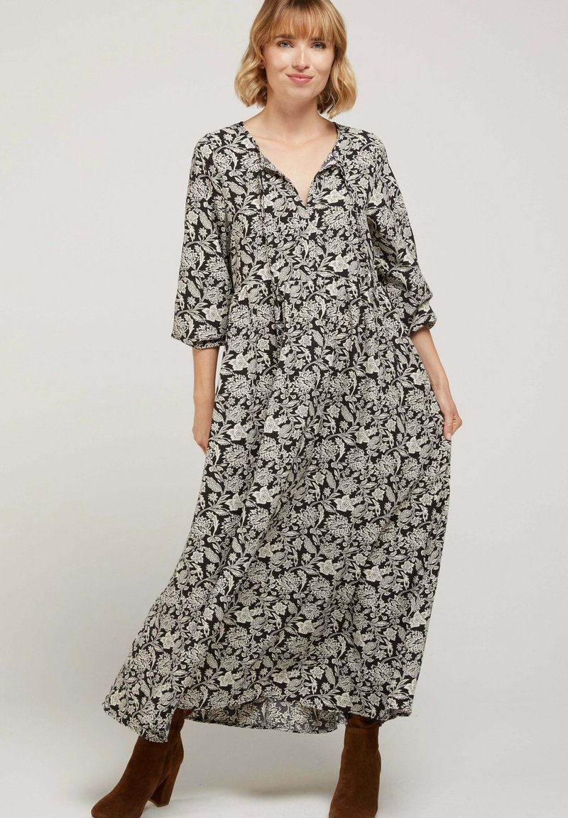 Robe maxi noire avec motif floral blanc, décolleté en V, manches trois-quarts et coupe loose, fabriquée en tissu léger. Bottes à talons brunes.